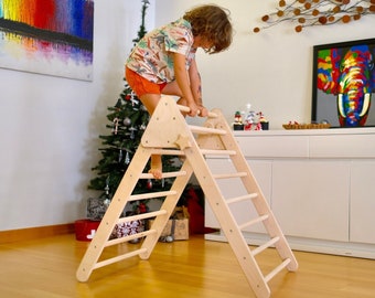 May include: A child climbs a wooden triangular climbing frame with a ladder and a rounded handrail. The frame is made of natural wood and has a light brown color. The child is wearing a floral shirt and orange shorts.