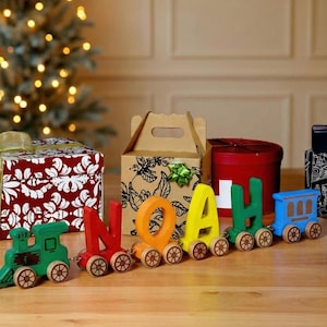 May include: A wooden toy train spelling out the name "NOAH" in colorful letters. The train is on a wooden surface, surrounded by wrapped gifts and a Christmas tree in the background. Festive holiday decor.