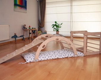 May include: A wooden climbing arch, slide, and cube set for toddlers. The natural wood pieces are connected and sit on a padded mat with a white background and gray star pattern. The set is designed for indoor play.