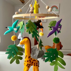 Può includere: Un mobile per bambini in legno a tema giungla. Presenta una girafa, una zebra, un leone, due uccelli e foglie verdi. Il mobile è sospeso da un anello di bambù.