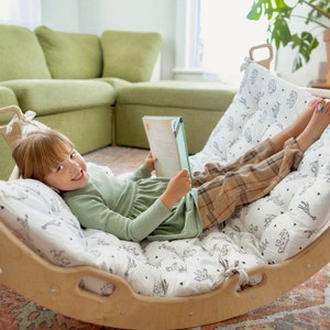 May include: A young person is lying in a wooden swing with a white and black patterned cushion. They are holding a book and smiling at the camera.