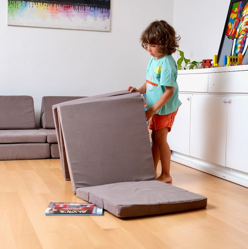 K&ouml;nnte beinhalten: Ein Kind baut eine graue Schaumstoff-Spielmatte zusammen. Die Matte besteht aus mehreren rechteckigen Teilen, die auf verschiedene Weise angeordnet werden k&ouml;nnen. Das Kind tr&auml;gt ein blaues Hemd und rote Shorts.