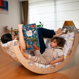 Può includere: Un genitore e un bambino stanno leggendo un libro insieme seduti su una sedia a dondolo di legno. Il libro è intitolato "Gute Nacht,  Schlummerbuch". La sedia a dondolo è rivestita di un tessuto a fantasia bianca e blu.