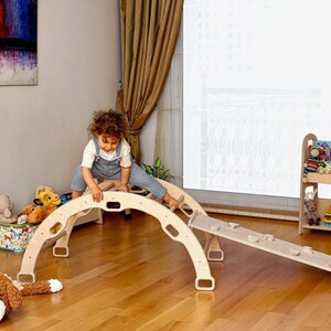 May include: A child plays on a wooden climbing arch and slide. The arch is made of natural wood and has a rounded top. The slide is attached to the arch and has a slight incline. The child is wearing a blue overall dress and has curly brown hair.
