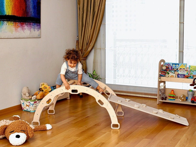 May include: A child plays on a wooden climbing arch and slide. The arch is made of natural wood and has a rounded top. The slide is attached to the arch and has a slight incline. The child is wearing a blue overall dress and has curly brown hair.
