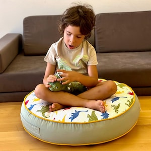 以下が含まれることがあります： 恐竜柄の丸い床クッションに座っている子供。クッションは薄い緑色で黄色の縁取りがされている。子供は緑色のぬいぐるみ恐竜を持っている。