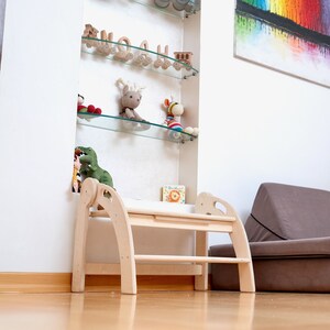 May include: A light wood step stool with two steps and rounded edges. The stool is in a room with a white wall and a glass shelf with toys on it.