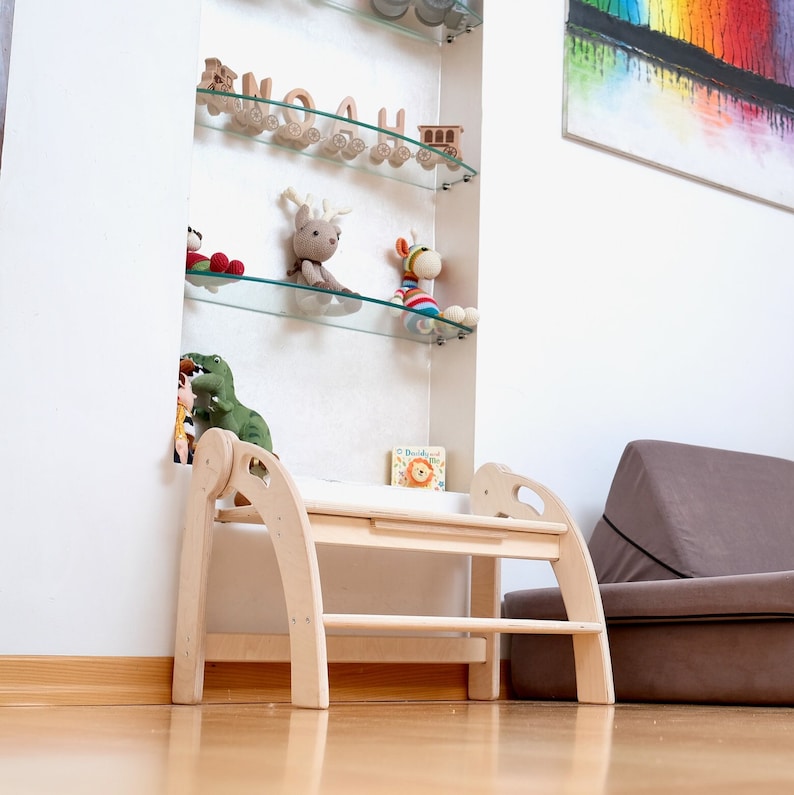 May include: A light wood step stool with two steps and rounded edges. The stool is in a room with a white wall and a glass shelf with toys on it.