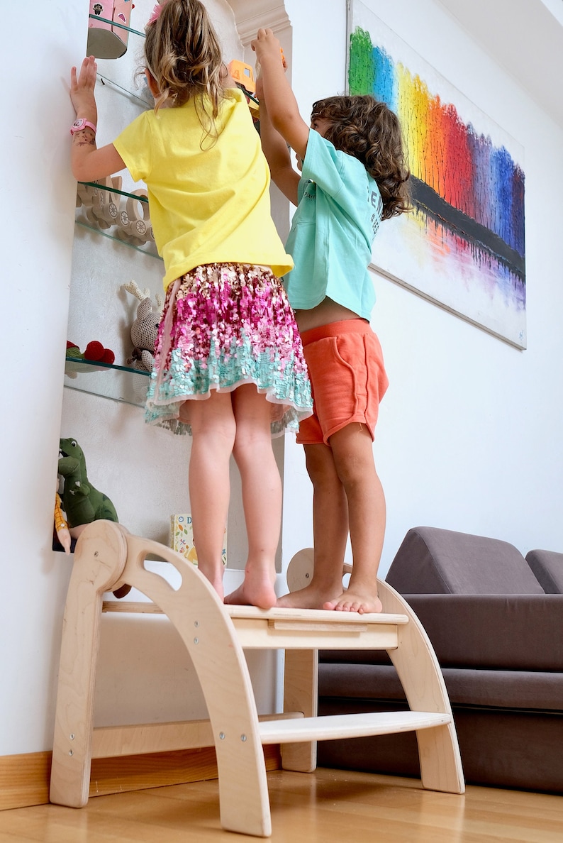 May include: Two children stand on a wooden step stool, reaching for items on a shelf. The girl is wearing a yellow shirt and a pink and green skirt. The boy is wearing a blue shirt and orange shorts. The step stool has two steps and is made of natural wood.