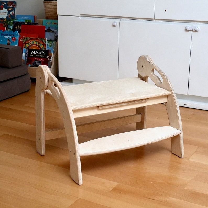 May include: A wooden step stool with a built-in table. The stool is made of light wood and has a curved design. The table is also made of light wood and has a small drawer.