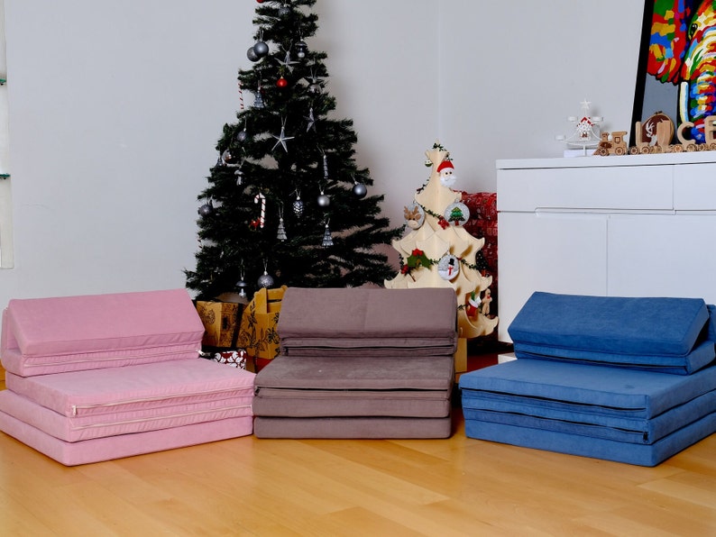 K&ouml;nnte beinhalten: Drei faltbare Futonmatratzen in Rosa, Braun und Blau. Die Matratzen sind zu einer Couchform gefaltet und stehen auf einem Holzboden. Im Hintergrund befinden sich Weihnachtsb&auml;ume und Dekorationen.