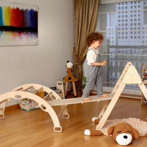 May include: A wooden playset for children, featuring a climbing triangle, a curved archway with rungs, and a wooden plank. A child is walking across the plank, and a fluffy brown dog rug is on the floor.