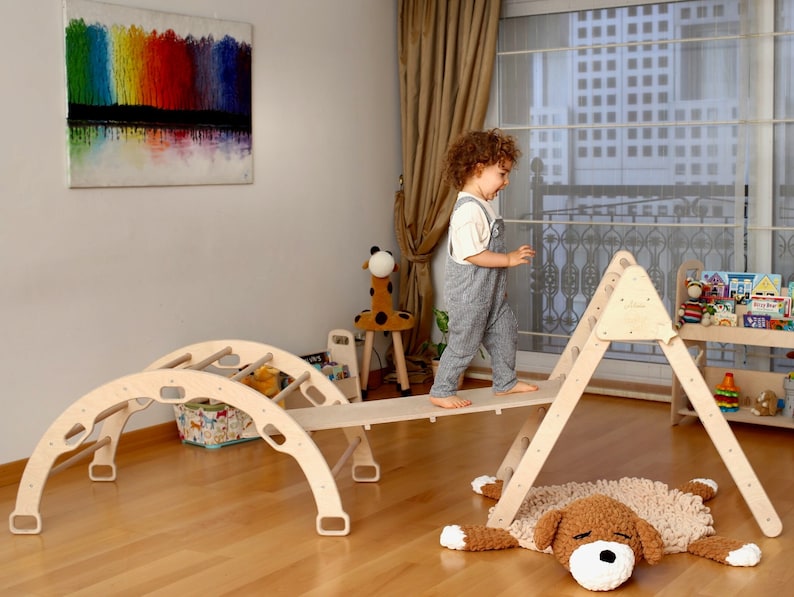 May include: A wooden playset for children, featuring a climbing triangle, a curved archway with rungs, and a wooden plank. A child is walking across the plank, and a fluffy brown dog rug is on the floor.