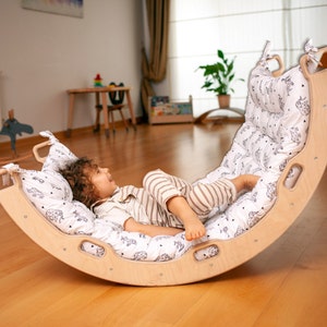 May include: A child lays on a wooden rocking balance board with a white and black patterned cushion. The board is curved and has a handle on each side. The child is wearing a striped shirt and pants.