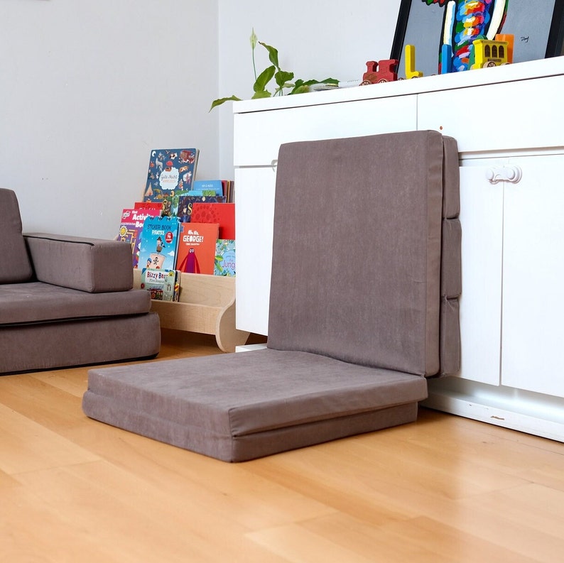 K&ouml;nnte beinhalten: Ein braunes, faltbares Bodenkissen mit dickem, gepolstertem Design. Das Kissen ist in der H&auml;lfte gefaltet und lehnt an einem wei&szlig;en Schrank. Das Kissen ist aus einem weichen, strukturierten Stoff.