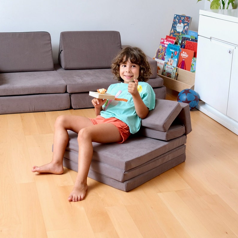 K&ouml;nnte beinhalten: Ein Kind sitzt auf einem grauen, zusammenklappbaren Futon-Sofa mit einem Snack in der Hand. Das Sofa besteht aus mehreren Schichten Schaumstoff und Stoff. Das Kind tr&auml;gt ein t&uuml;rkisfarbenes T-Shirt und rote Shorts.