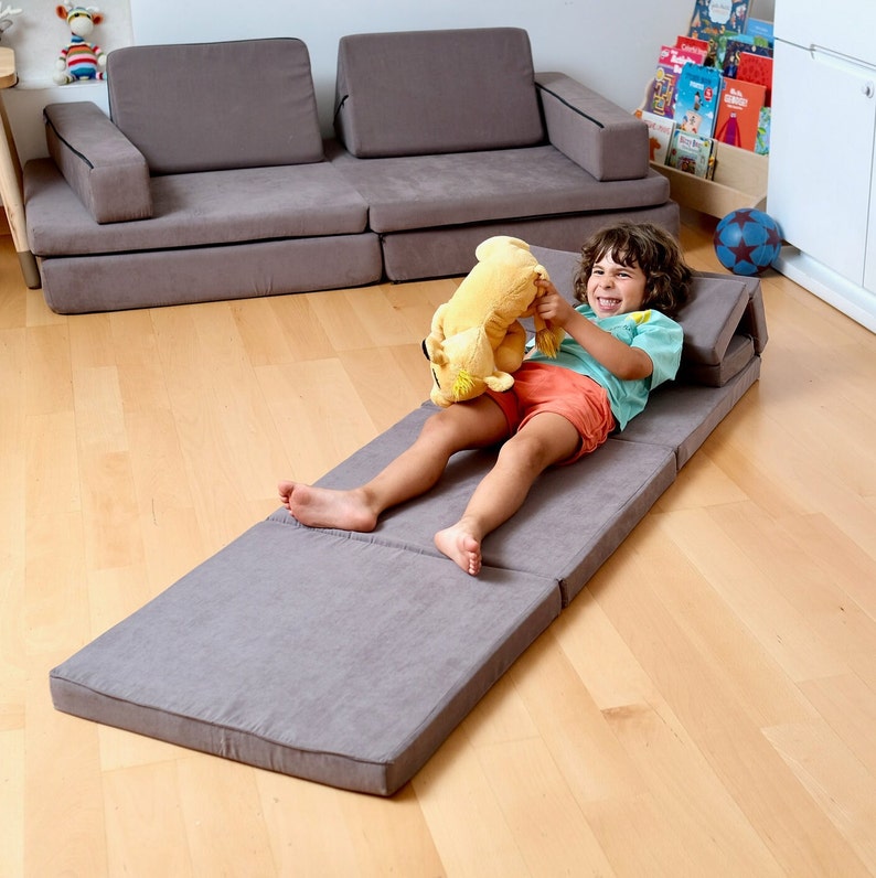K&ouml;nnte beinhalten: Eine graue, zusammenklappbare Futonmatratze, auf der ein Kind liegt. Das Kind h&auml;lt ein gelbes Stofftier und l&auml;chelt. Der Futon ist ausgeklappt und hat einen grauen Stoffbezug.