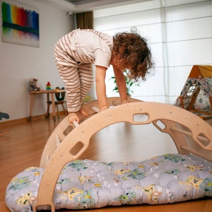 以下が含まれることがあります： 子供が、パッド付きのベースが付いた木製のアーチを登っています。アーチには梯子と丸い開口部があります。子供はストライプのパンツと白いシャツを着ています。