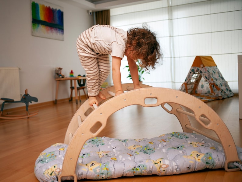 以下が含まれることがあります： 子供が、パッド付きのベースが付いた木製のアーチを登っています。アーチには梯子と丸い開口部があります。子供はストライプのパンツと白いシャツを着ています。