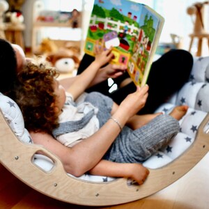 May include: A wooden rocking chair with a white and gray star patterned cushion. A parent and child are sitting in the chair reading a book together.
