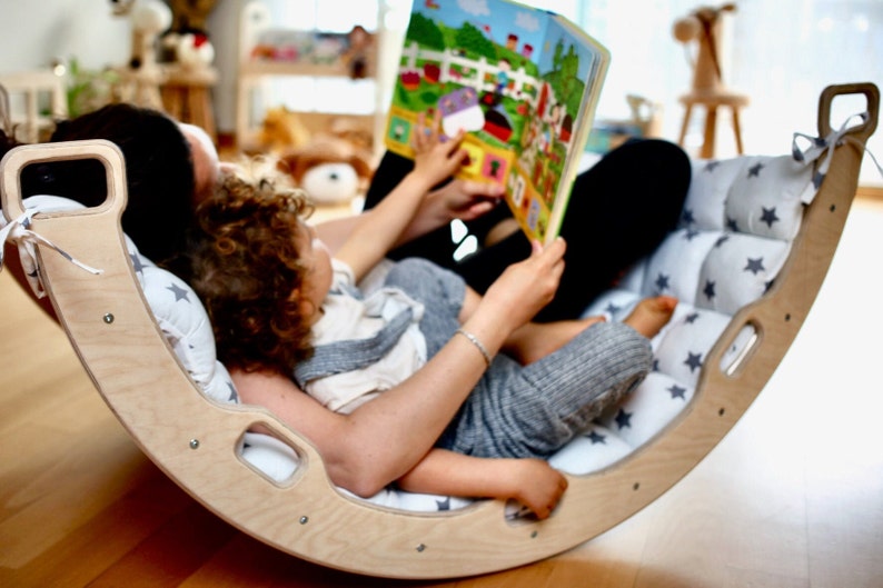 May include: A wooden rocking chair with a white and gray star patterned cushion. A parent and child are sitting in the chair reading a book together.