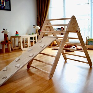 May include: A wooden triangular climbing frame with a slide attached. The frame has a ladder with rungs and the slide has a textured surface with small wooden shapes. The frame is made of natural wood and is suitable for children's play.
