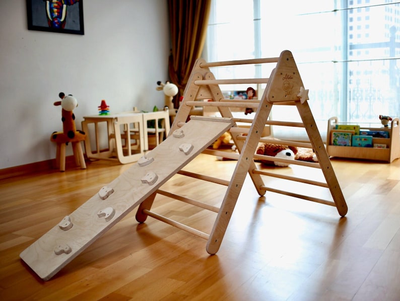 May include: A wooden triangular climbing frame with a slide attached. The frame has a ladder with rungs and the slide has a textured surface with small wooden shapes. The frame is made of natural wood and is suitable for children's play.