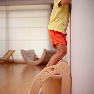 May include: A young child stands on a wooden step stool, reaching up to touch a white wall. The stool has two steps and a rounded top. The child is wearing a yellow shirt and orange shorts.