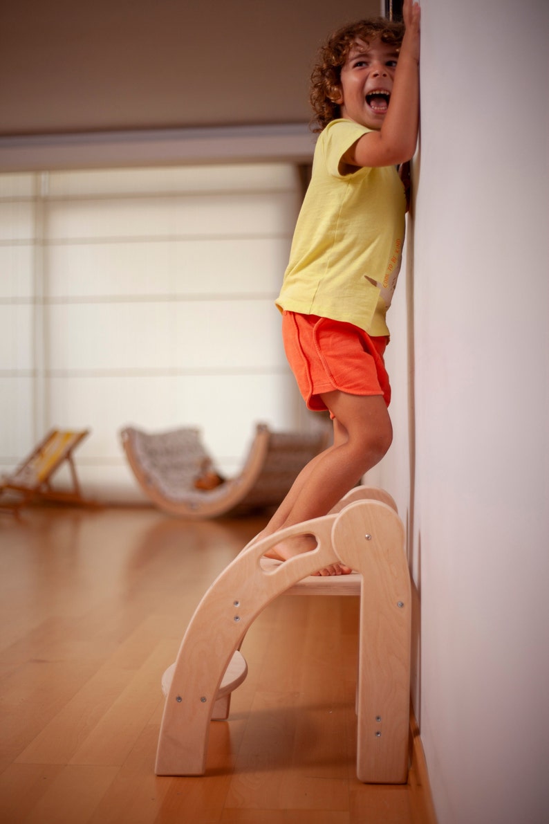 May include: A young child stands on a wooden step stool, reaching up to touch a white wall. The stool has two steps and a rounded top. The child is wearing a yellow shirt and orange shorts.