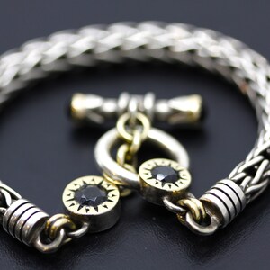 May include: A silver bracelet with a braided chain design and a gold toggle clasp. The clasp features two black gemstones.