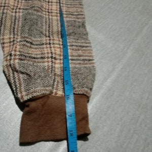 May include: Close-up of a brown and beige plaid fabric with a brown cuff. A blue measuring tape is visible along the right edge of the image, with the numbers 14 to 20 inches visible.