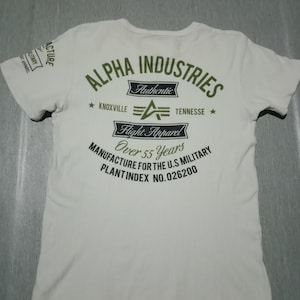 Könnte beinhalten: Weißes T-Shirt mit einem grünen Aufdruck des Alpha Industries Logos. Das Logo enthält den Text "Alpha Industries", "Authentic", "Flight Apparel", "Knoxville", "Tennessee", "Over 55 Years", "Manufacture for the U.S. Military" und "Plantindex No. 026200".