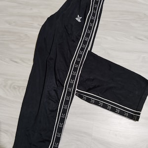May include: Black sweatpants with white side stripes featuring a repeated logo design. The logo is a stylized bird with wings outstretched.