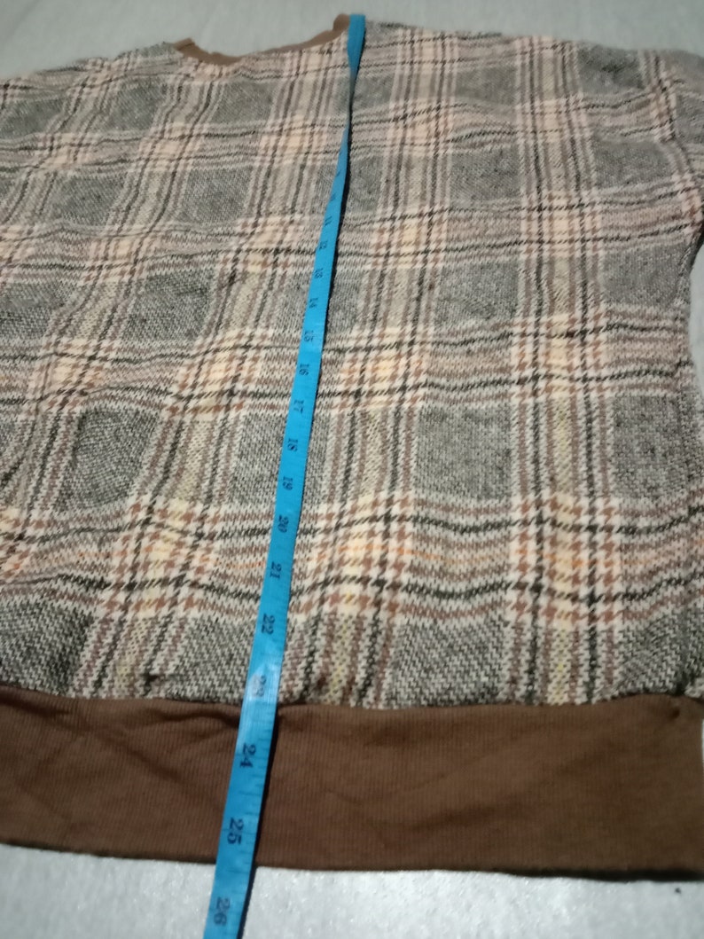 May include: Brown and beige plaid sweater with a brown ribbed hem. The sweater is measured with a blue tape measure.