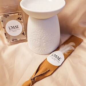 May include: A white ceramic essential oil diffuser with a tree of life design, a wax melt, and a wrapped feather. The wax melt is in a clear plastic container and has brown specks. The wrapped feather has a label that says "L'MAE".