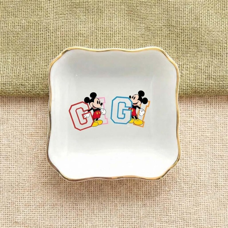 May include: White square trinket dish with a gold rim. The dish features two Mickey Mouse illustrations, each next to a letter "G" in red and blue. The background is a textured, light brown fabric.
