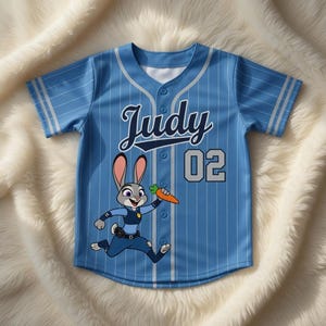 Puede incluir: Camiseta de b&eacute;isbol azul claro con rayas blancas, con el nombre "Judy" y el n&uacute;mero "02". Una imagen de dibujos animados de un conejo sosteniendo una zanahoria est&aacute; en la parte delantera. La camiseta tiene una parte delantera abotonada y ribetes blancos en las mangas.