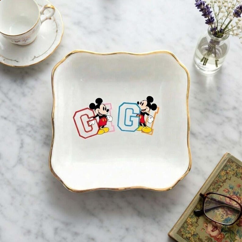 May include: A white square decorative tray with a gold rim. The tray features two Mickey Mouse illustrations, each next to a letter "G" in a collegiate style. One "G" is red, the other is blue.