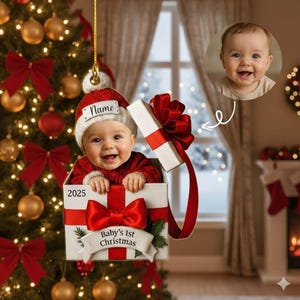 May include: A Christmas ornament featuring a baby in a gift box with a red bow. The baby wears a red sweater and a Santa hat with the name "Name" on it. The ornament also has the text "Baby's 1st Christmas" and the year "2025".