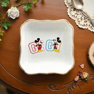May include: White square ceramic dish with a gold rim, featuring two Mickey Mouse figures flanking the letter "G" in red and blue. The dish is on a wooden surface, with floral accents and a spoon visible.