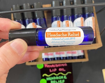 May include: A blue glass roll-on bottle labeled "Headache Relief" with a black cap. The label includes the text "GoldenAgeMassage.com" and details about the oil's ingredients. Several similar bottles are visible in the background.