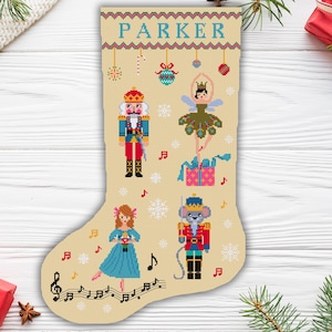 Op de afbeelding: Een beige cross-stitch kousenpatroon met een Notenkraker, een ballerina, een muis en een cadeaudoos. De kous is versierd met sneeuwvlokken, muzieknoten en de naam "PARKER".