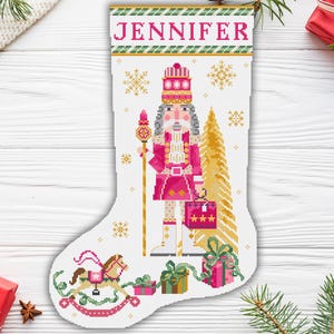 Pode incluir: Uma meia de Natal rosa e branca com um design de Quebra-Nozes. A meia tem uma borda verde e dourada com o nome "Jennifer" no topo. O Quebra-Nozes está segurando um cajado e uma caixa de presente. Há um cavalinho de balanço e uma árvore de Natal na parte inferior da meia.