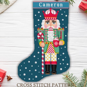 May include: Cross stitch pattern for a Christmas stocking featuring a nutcracker holding a cup of hot chocolate. The stocking is blue with white snowflakes and the nutcracker is wearing a green and red uniform with a candy cane in his hand. The stocking is personalised with the name "Cameron".
