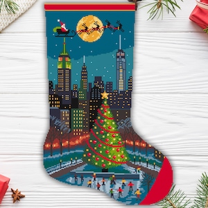 Op de afbeelding: Een kerstsok met een kruissteekpatroon met de skyline van New York City 's nachts. De kerstman vliegt over de stad. Een versierde kerstboom staat op de voorgrond en de tekst "CROSS STITCH PATTERN" staat onderaan.
