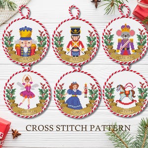 Peut inclure: Six ornements de Noël ronds en point de croix avec des cordes rouges et blanches. Chaque ornement représente un personnage différent : Casse-Noisette, Soldat de plomb, Roi des souris, Sucre d'orge, Clara et un cheval à bascule. Le texte "Cross Stitch Pattern" est en bas.