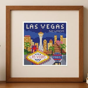Könnte beinhalten: Ein gerahmtes Kreuzstich-Kunstwerk, das die Skyline von Las Vegas zeigt, einschließlich des Stratosphere Tower und des "Welcome to Las Vegas"-Schildes. Das Kunstwerk befindet sich in einem Holzrahmen und steht auf einer Holzoberfläche.