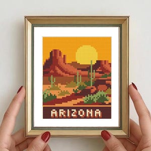 Peut inclure: Broderie au point de croix d'un paysage désertique de l'Arizona, avec un coucher de soleil, des cactus et des formations rocheuses rouges. Le mot "ARIZONA" est brodé en bas. L'œuvre est encadrée dans un cadre doré.