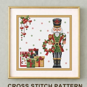 May include: A framed cross stitch pattern featuring a nutcracker in a green and red uniform holding a wreath and candy cane, surrounded by wrapped gifts, holly, and small peppermint candies on a white grid background with a gold border.