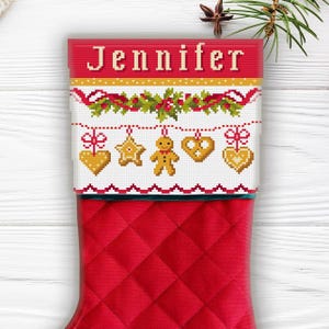 Könnte beinhalten: Roter Weihnachtsstrumpf mit einem Steppmuster. Die Oberseite zeigt ein Kreuzstichdesign mit dem Namen "Jennifer" und festlichen Lebkuchenmännchen-, Stern- und Herzornamenten. Eine grüne Bordüre akzentuiert den oberen Rand.